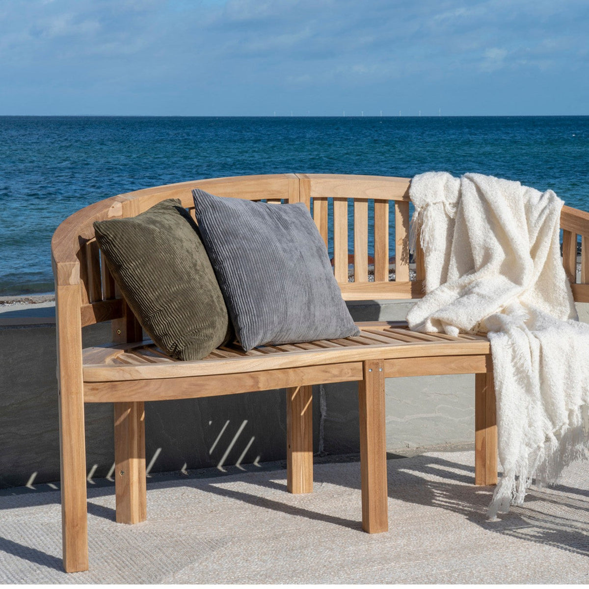 Havemøbler i teak: buet havebænk med puder og et lunt plaid på en terrasse ved havet