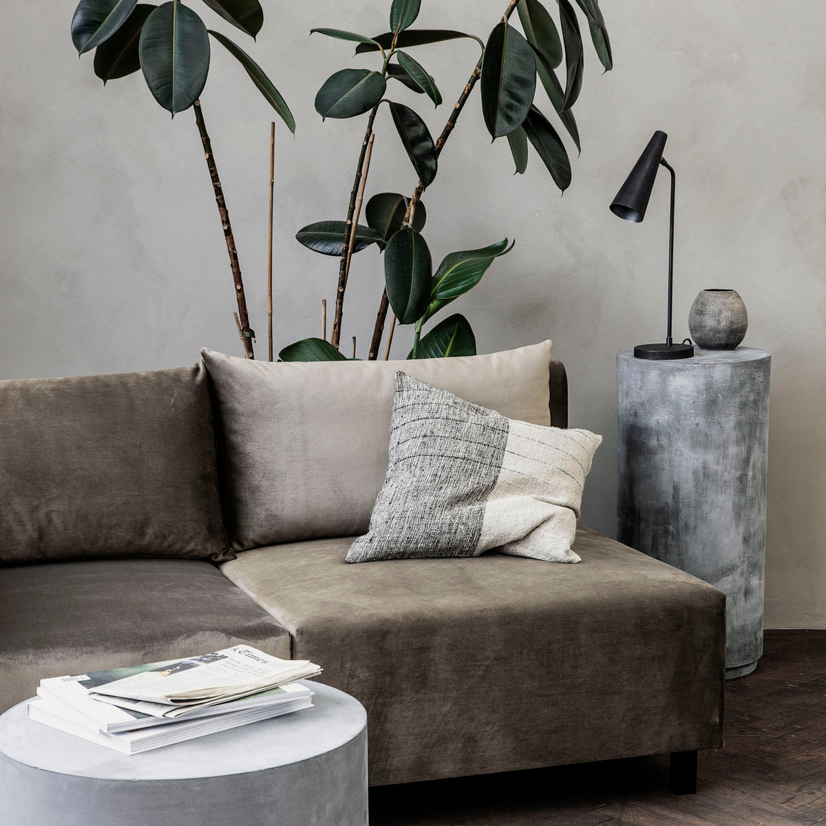 Indretning af stue med veloursofa, pyntepuder, stort gummitræ og sort bordlampe på rundt sidebord