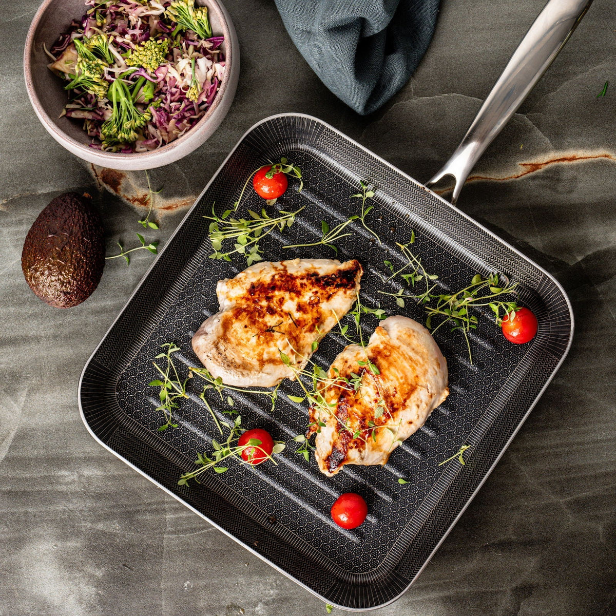 Grillpande med stegt kyllingebryst, cherrytomater og krydderurter set fra oven på en mørk stenbordplade