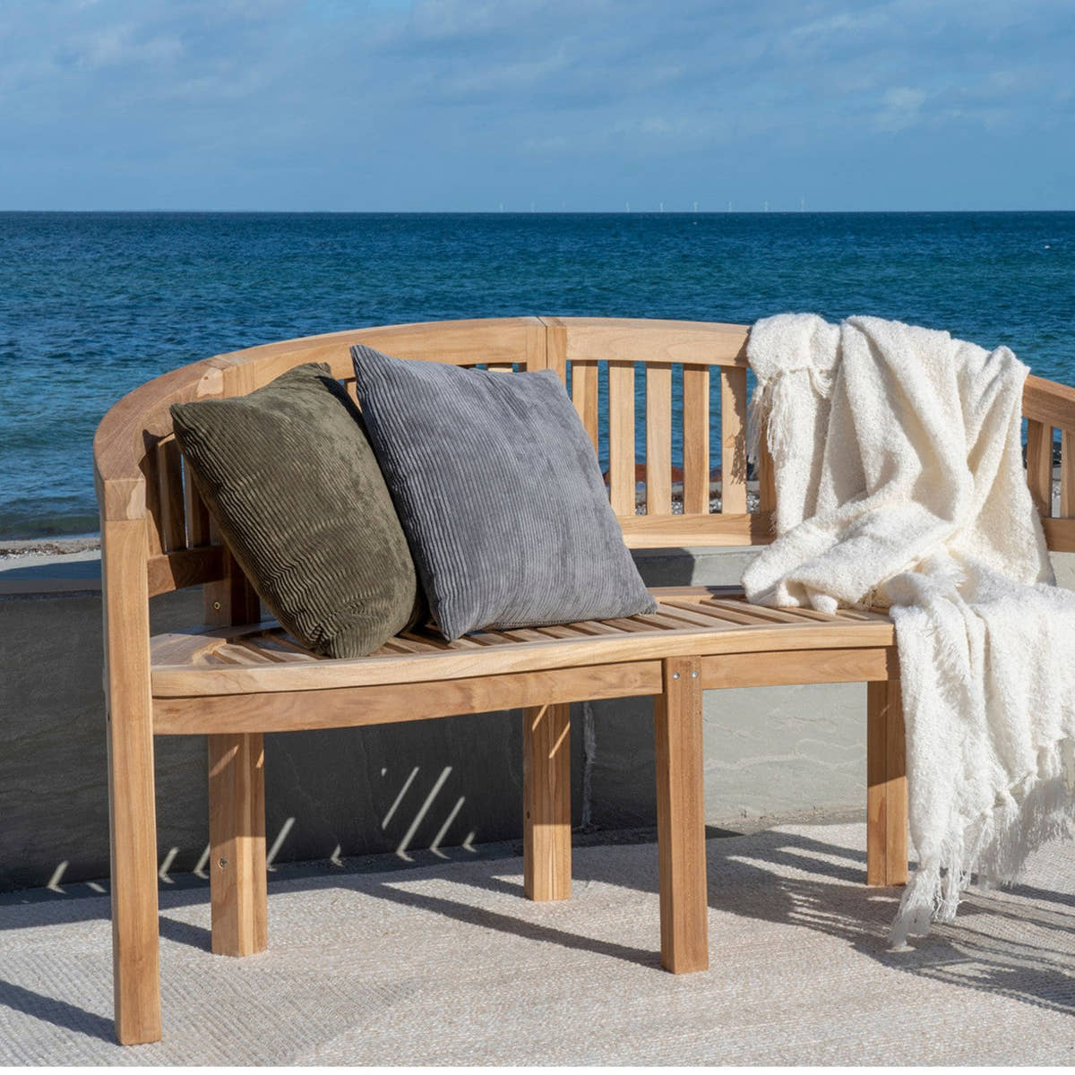 Havemøbler i teak: buet havebænk med puder og et lunt plaid på en terrasse ved havet